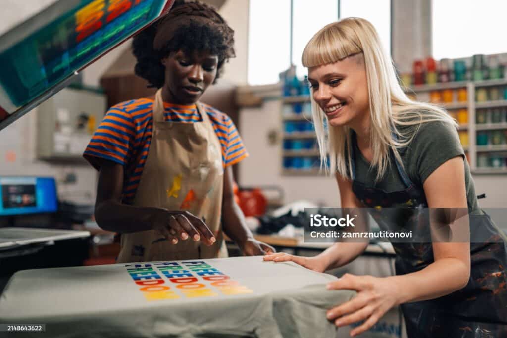 Smiling multicultural female printing workshop technicians standing at printing workshop and silkscreen printing textile products and t-shirt while promoting lgbtq equality.Females in graphic industry